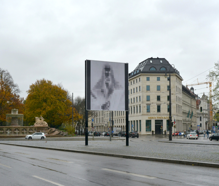 Diagonal view of the billboard at Lenbachplatz. The motif shows a black-and-white reproduction of Albrecht Dürer's "Self-Portrait in a Fur Coat" in negative, pixelated in large squares.