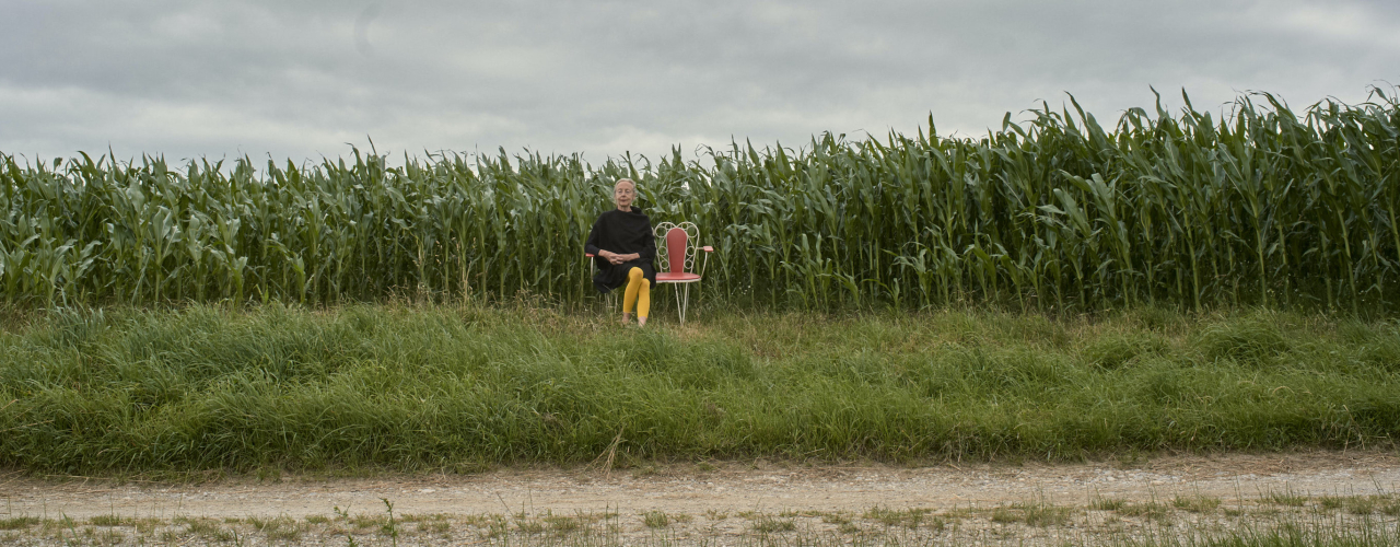 In the background is a cornfield against a cloudy sky. In front of it is a white metal bench with red cushions, on which sits a blonde woman in a black dress and yellow tights.