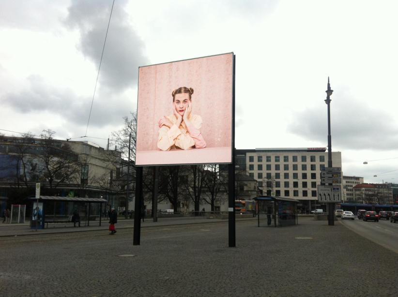View of the billboard on Lenbachplatz looking towards the city with the motif "Dreiarmige Frau" by Susanne Steinmaßl. The motif shows a blonde woman in a pale pink blouse sitting in front of a pink patterned wallpaper. She has two arms propped up on a pink table in front of her and rests her face in both hands. The woman also has a third arm, which she is resting on the table and clasping her shoulder with her hand.