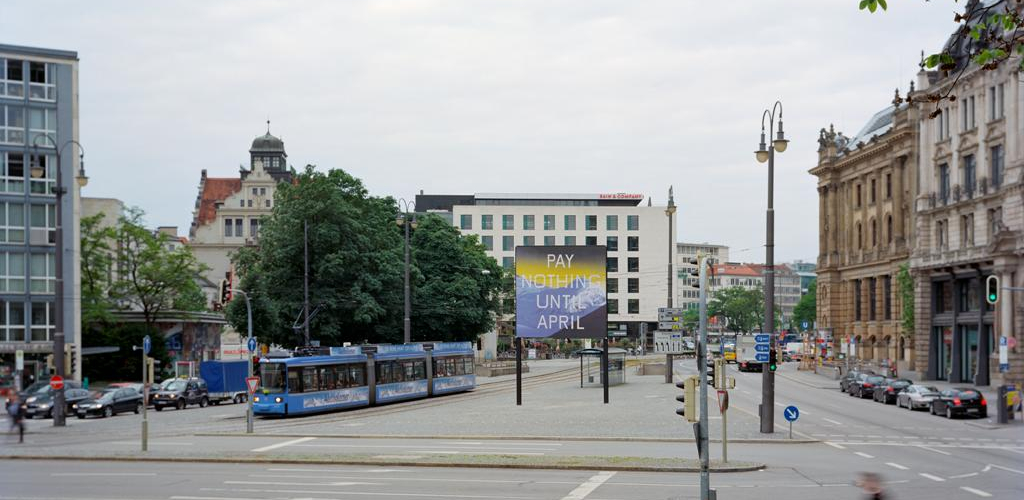 Frontal view of Lenbachplatz looking towards the city center. The billboard is placed in the middle of the square, with a streetcar running alongside. The billboard features a work by the artist Ed Ruscha. The motif shows a snow-covered mountain range in blue with a yellowish horizon above it. The inscription "Pay Nothing Until April" appears on the picture in white capital letters.