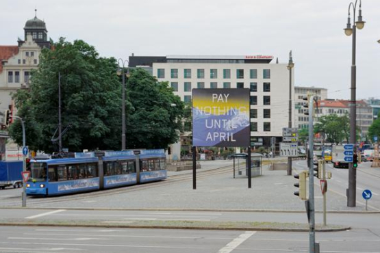 Frontal view of Lenbachplatz looking towards the city center. The billboard is placed in the middle of the square, with a streetcar running alongside. The billboard features a work by the artist Ed Ruscha. The motif shows a snow-covered mountain range in blue with a yellowish horizon above it. The inscription "Pay Nothing Until April" appears on the picture in white capital letters.