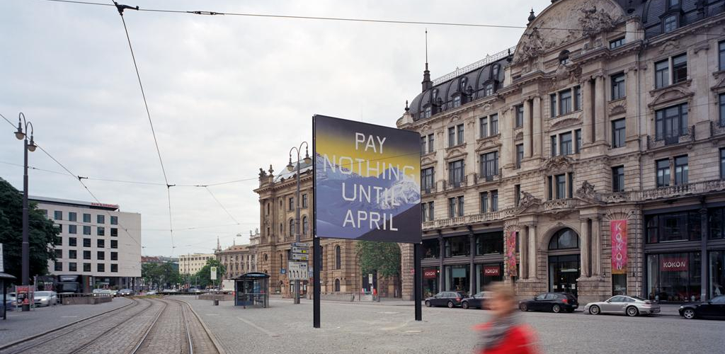 Seitliche Ansicht des Billboards am Lenbachplatz mit Blick Richtung Innenstadt. Auf dem Billboard zu sehen ist eine Arbeit des Künstlers Ed Ruscha. Das Motiv zeigt eine in blau gehaltene verschneite Bergkette und darüber ein gelblicher Horizont. Auf dem Bild erscheint die Aufschrift: "Pay Nothing Until April" in weißen Großbuchstaben. Im Vordergrund läuft eine Frau in rotem Mantel vorbei.