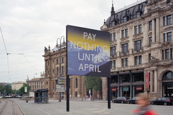 Seitliche Ansicht des Billboards am Lenbachplatz mit Blick Richtung Innenstadt. Auf dem Billboard zu sehen ist eine Arbeit des Künstlers Ed Ruscha. Das Motiv zeigt eine in blau gehaltene verschneite Bergkette und darüber ein gelblicher Horizont. Auf dem Bild erscheint die Aufschrift: "Pay Nothing Until April" in weißen Großbuchstaben. Im Vordergrund läuft eine Frau in rotem Mantel vorbei.