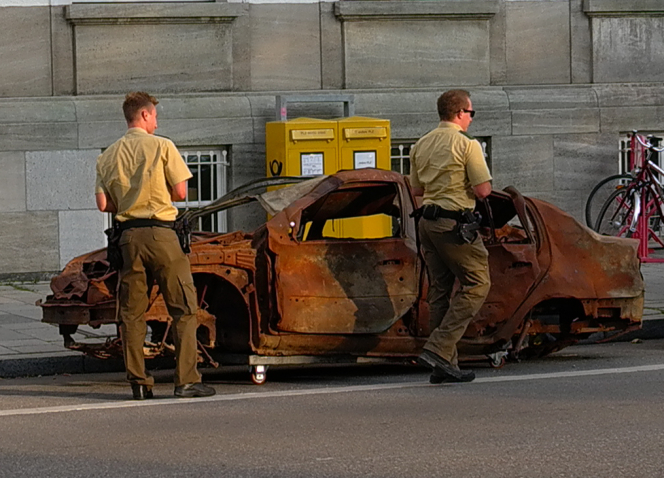 Fotografie eines braunen ausgebrannten Autowracks ohne Reifen, das auf der Maximilianstraße in München geparkt ist. Vor dem Wrack stehen zwei Polizisten, die um das Auto herumgehen und es inspizieren.