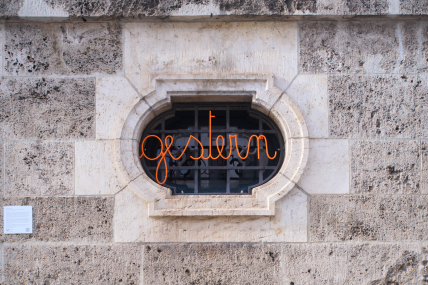 Zu sehen ist eine Hauswand mit einem Fenstergitter. In diesem Fenstergitter befindet sich ein Hakenkreuz-Muster, davor der geschwungene orangefarbene Schriftzug 'gestern'.