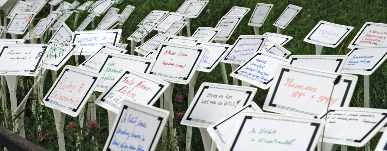 Fotografie einer Wiese im Hofgarten, in die viele weiße Schilder gesteckt sind. Auf die Schilder sind in unterschiedlichen Farben Begriffe und Sätze geschrieben.