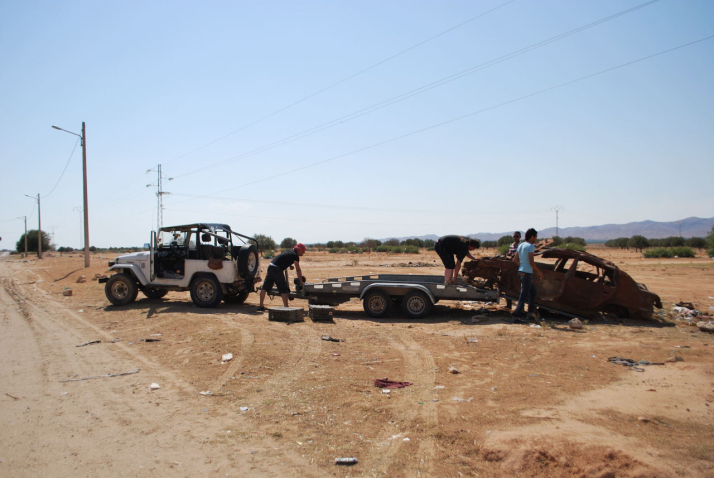 Bild eines ausgebrannten Autowracks am Rande einer Schotterstraße in Tunesien. Das Autowrack wird von vier Männern auf einen Anhänger verladen, der von einem weißen Geländewagen gezogen wird.