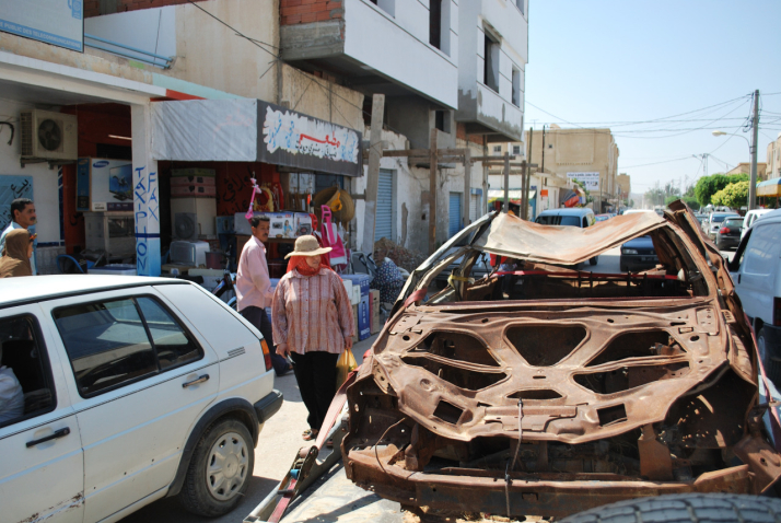 Bild eines ausgebrannten braunen Autowracks, das auf einen Autoanhänger geschnallt ist und durch eine Straßen von Sidi Bouzid, Tunesien, gezogen wird. Neben dem Autowrack sind geparkte und vorbeifahrende Autos zu sehen, zudem Passant*innen, die die Szene betrachten.