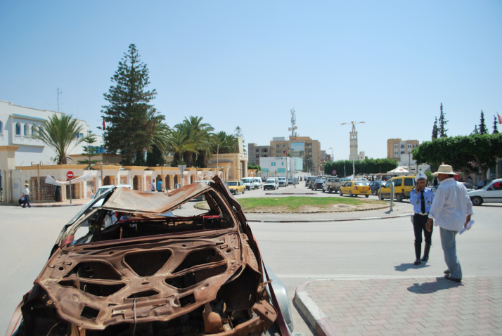 Bild eines ausgebrannten braunen Autowracks, das am Rand einer Straße in Sidi Bouzid, Tunesien, geparkt ist. Im Hintergrund zu sehen ist eine befahrene Straße mit einem Kreisverkehr sowie höhere Häuser.