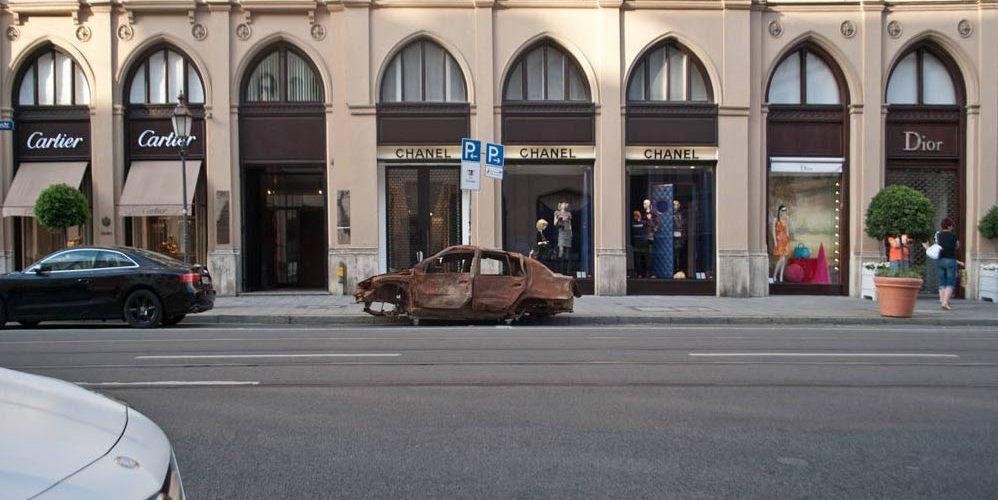 Fotografie eines braunen ausgebrannten Autowracks ohne Reifen, das auf der Maximilianstraße in München vor dem Chanel Shop geparkt ist.