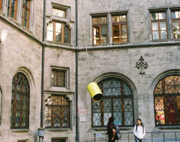 View of the inner courtyard of the Munich City Hall. A yellow-painted metal barrel hangs in the air between several ropes attached to the walls of the building. The barrel, which is open at the bottom, is a contemporary installation by artist Florian Froese-Peeck entitled 'Freisprechanlage'.
