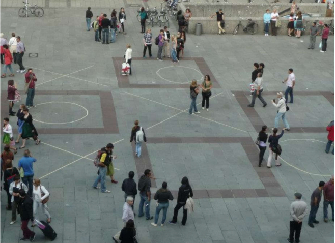 View of Marienplatz from above. There are many passers-by and tourists. Large crosses and circles are painted on the ground of the square, just like in the game 'Tic-Tac-Toe'.