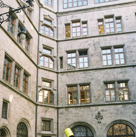 View of the inner courtyard of the Munich City Hall. A yellow-painted metal barrel hangs in the air between several ropes attached to the walls of the building. The barrel, which is open at the bottom, is a contemporary installation by artist Florian Froese-Peeck entitled 'Freisprechanlage'.