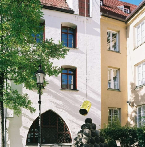 View of the inner courtyard of the Münchner Stadtmuseum. Above the sculpture of the stone spherical pyramid, a yellow-painted metal barrel hangs in the air, suspended between several ropes. The barrel, which is open at the bottom, is a contemporary installation by the artist Florian Froese-Peeck entitled "Freisprechanlage".