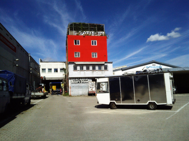 Photograph of the Box Factory site. A multi-storey red and grey building can be seen in the centre, identified as the "Boxfabrik" by two signs on the building. In the background are some industrial buildings. An old white and black van is parked in the forecourt in front of the box factory.