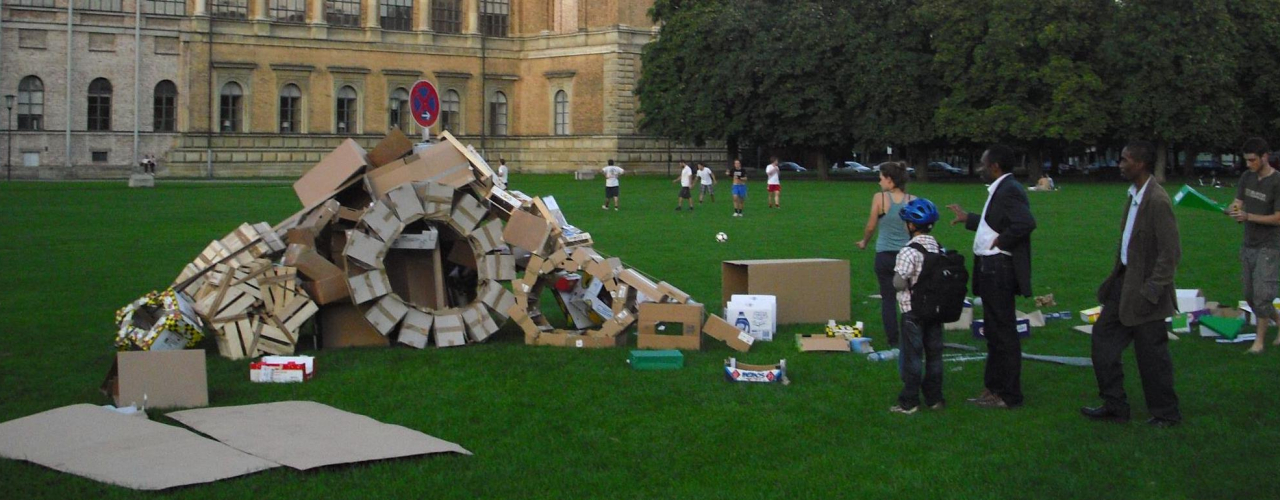Photograph of an installation on the lawn behind the Alte Pinakothek. The installation by artist Valerie Christiansen consists of various old packaging materials, mainly cardboard boxes and plywood fruit crates. The various boxes are arranged to form wheels and a kind of ramp, with a "no parking" sign in the middle. A few passers-by stand around the installation and look at it.