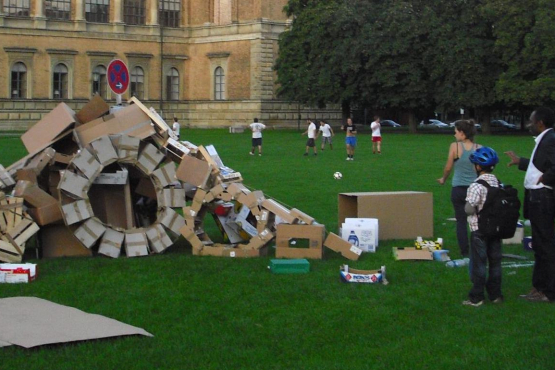 Photograph of an installation on the lawn behind the Alte Pinakothek. The installation by artist Valerie Christiansen consists of various old packaging materials, mainly cardboard boxes and plywood fruit crates. The various boxes are arranged to form wheels and a kind of ramp, with a "no parking" sign in the middle. A few passers-by stand around the installation and look at it.