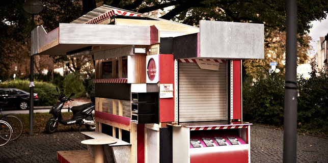 Photography of a kiosk-like installation in a public space. The canopied kiosk, in black, red and white, is made of wooden panels and its sides are designed differently. The front has several compartments with a bench and a table in front of them. Another side has a hatch with lowered blinds and a display compartment for flyers underneath.