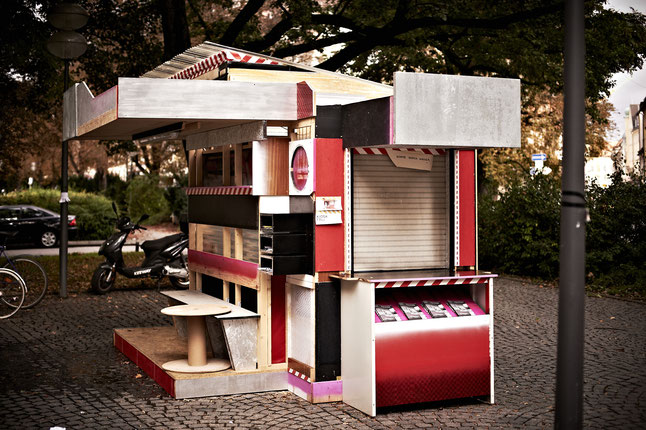 Photography of a kiosk-like installation in a public space. The canopied kiosk, in black, red and white, is made of wooden panels and its sides are designed differently. The front has several compartments with a bench and a table in front of them. Another side has a hatch with lowered blinds and a display compartment for flyers underneath.