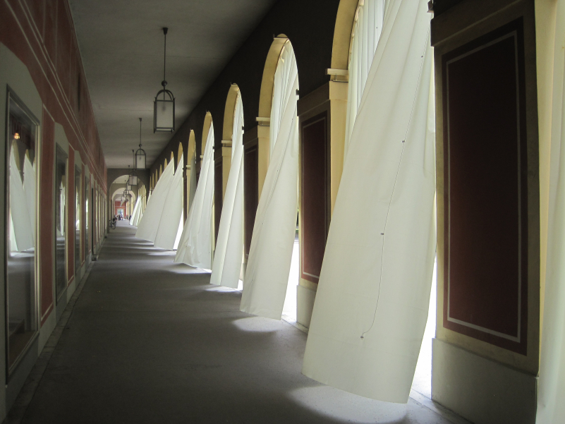 Fotografie in die Hofgartenarkaden. Die Arkadenbögen sind mit weißen Vorhängen verhangen, die im Wind flattern.