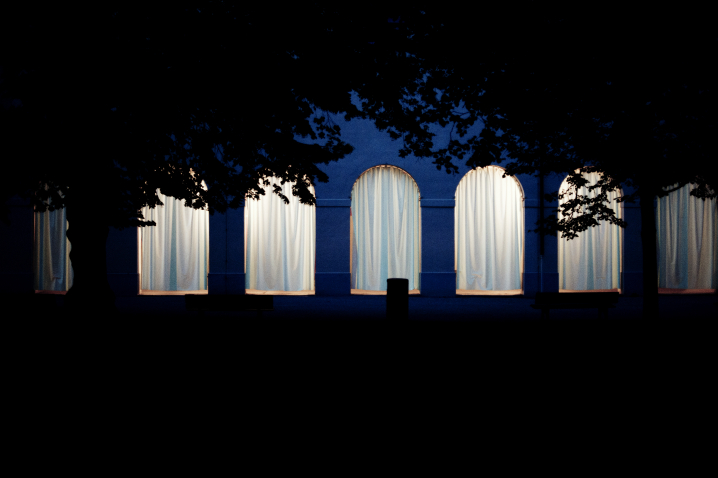 Frontale Ansicht der Hofgartenarkaden bei Nacht. Die Arkadenbögen sind mit weißen Vorhängen verhangen, die dahinterliegenden Arkadengänge sind erleuchtet und das Licht scheint durch die Vorhänge hindurch.