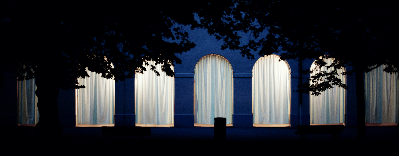 Frontale Ansicht der Hofgartenarkaden bei Nacht. Die Arkadenbögen sind mit weißen Vorhängen verhangen, die dahinterliegenden Arkadengänge sind erleuchtet und das Licht scheint durch die Vorhänge hindurch.