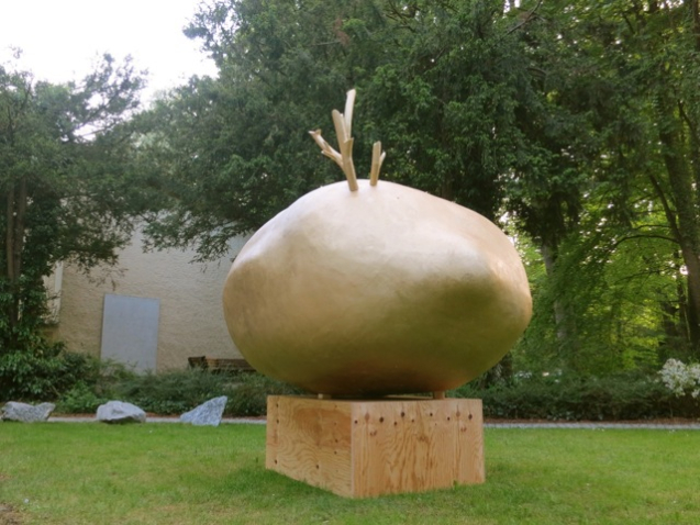 Photograph of a park, an installation by the artist Alix Stadtbäumer stands in a green space. The installation consists of a wooden pedestal on which rests a golden sculpture of a potato with golden sprouts.