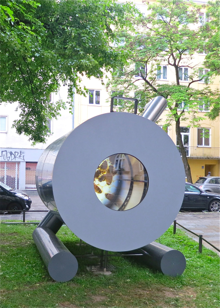 Photography of a silver stove sculpture by the artists Alix Stadtbäumer and Yvonne Leinfelder. This is placed on the lawn on the lawn at Rumford-Ecke-Klenzestraße. A video work by Yvonne Leinfelder can be seen through a kind of porthole inside the artwork. The sculpture refers to one of Count Rumford's most important inventions, the Rumford stove.