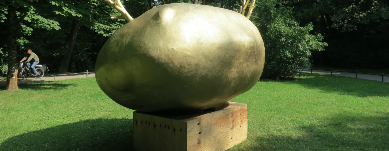 Photograph of a green space in the English Garden, where an installation by artist Alix Stadtbäumer has been placed. The installation consists of a wooden plinth on which rests a gold-coloured sculpture of a potato from which golden shoots are sprouting.
