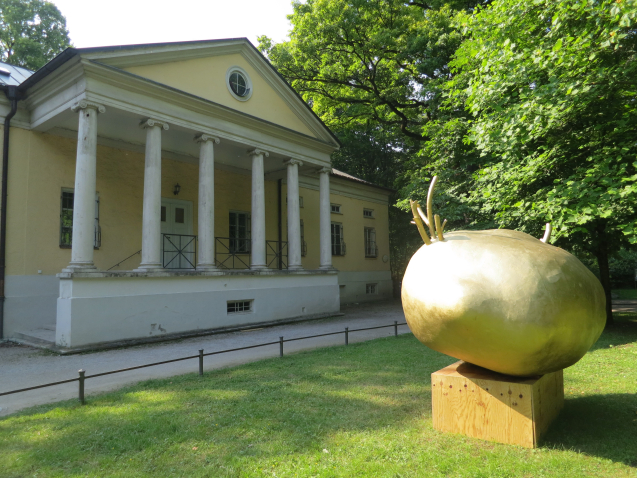 Photograph of a green space in the English Garden, on which an installation by the artist Alix Stadtbäumer has been placed. The installation consists of a wooden pedestal on which rests a golden sculpture of a potato with golden sprouts. In the background is a classicistic building with a colonnaded portico, the so called Rumfordschlössl.