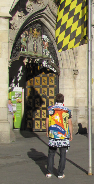 Photograph of a man standing on Marienplatz in front of the New Town Hall. He has his back to the viewer and is standing next to a flagpole on which the black and yellow Munich flag is hoisted. He is wearing a sleeveless, colourful waistcoat that appears to be a collage of different pieces of fabric. The pieces of fabric are textile items from various Munich clubs, which have been put together to form a patchwork-like 'Munich Club Collage' in the form of a garment.