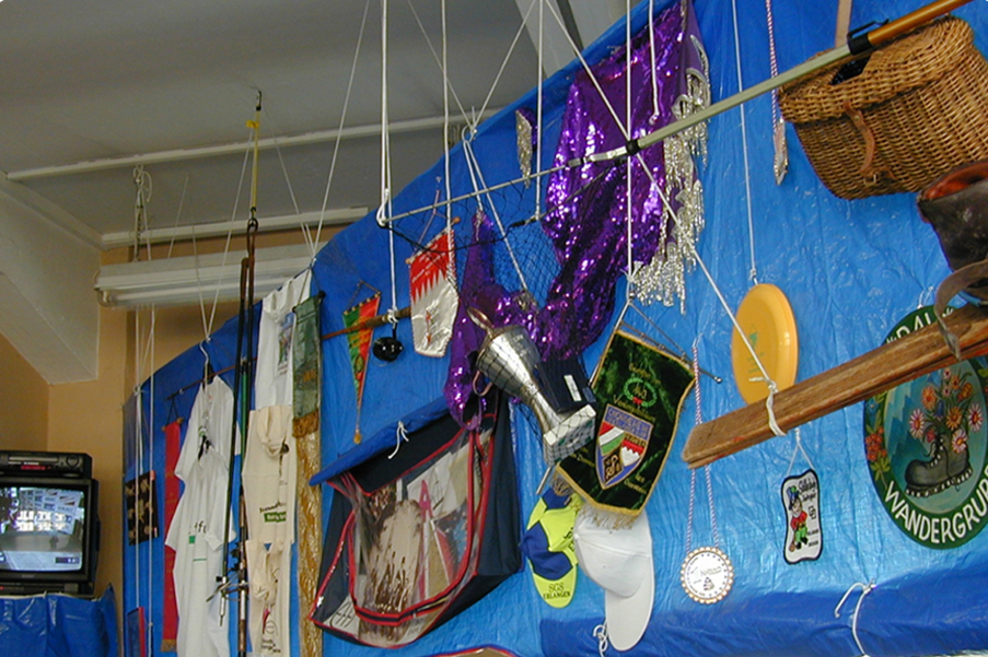 Photograph of a room with a ceiling installation. A large rectangular blue plastic sheet hangs from the ceiling. Various objects from different Munich clubs are attached to this tarpaulin. There are various pennants, t-shirts, baseball caps, trophies, costumes and other club paraphernalia.