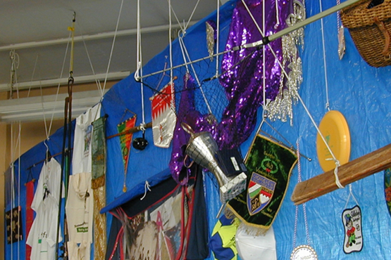 Photograph of a room with a ceiling installation. A large rectangular blue plastic sheet hangs from the ceiling. Various objects from different Munich clubs are attached to this tarpaulin. There are various pennants, t-shirts, baseball caps, trophies, costumes and other club paraphernalia.