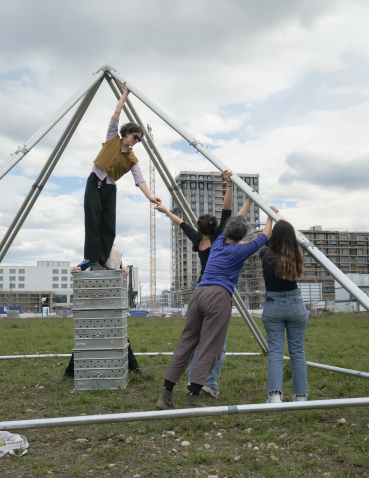 Vier Frauen bauen ein pyramidenförmiges Gerüst auf. Im Hintergrund sieht man die Großbaustelle Freiham.