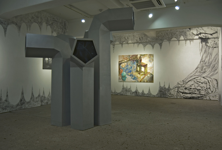 View of an exhibition in a windowless room. In the foreground is an installation of five grey objects of varying sizes that resemble giant ventilation pipes. In the background, two paintings hang on the walls. The walls themselves are also painted in black and white with various motifs, some of which are reminiscent of an abstract stalactite cave.