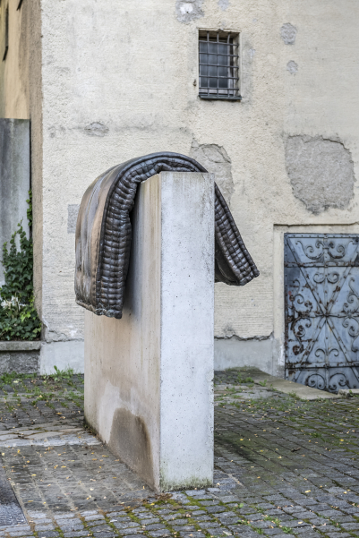 Bild einer Brunnenskulptur von Tatiana Trouvé vor der Mauer des alten Südfriedhofs am Stephansplatz. Die Skulptur besteht aus einem Brozeguss einer Matratze, die über einen Betonsockel gelegt ist.