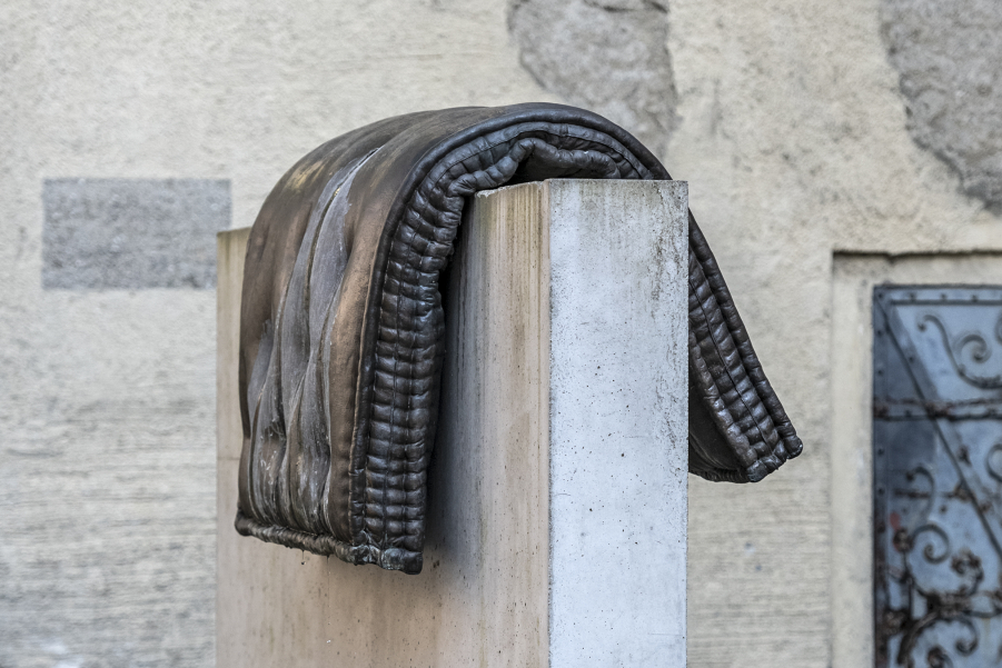 Image of a fountain sculpture by Tatiana Trouvé in front of the wall of the old South Cemetery on Stephansplatz. The sculpture consists of a bronze cast of a mattress placed over a concrete base.