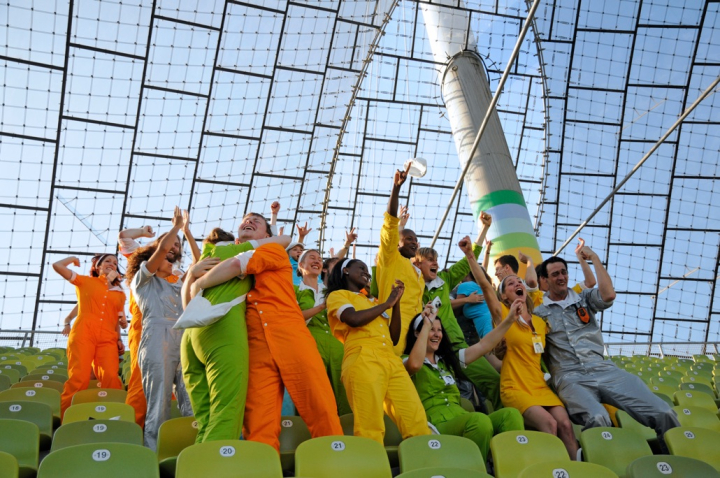 The picture shows a photograph taken in the Olympic Stadium. A group of performers stand in the green rows of chairs under the tent roof, joyfully and jubilantly raising their arms in the air, clapping their hands or lying in each other's arms. Each performer wears a monochrome jumpsuit in colours ranging from orange and green to blue, silver and yellow.