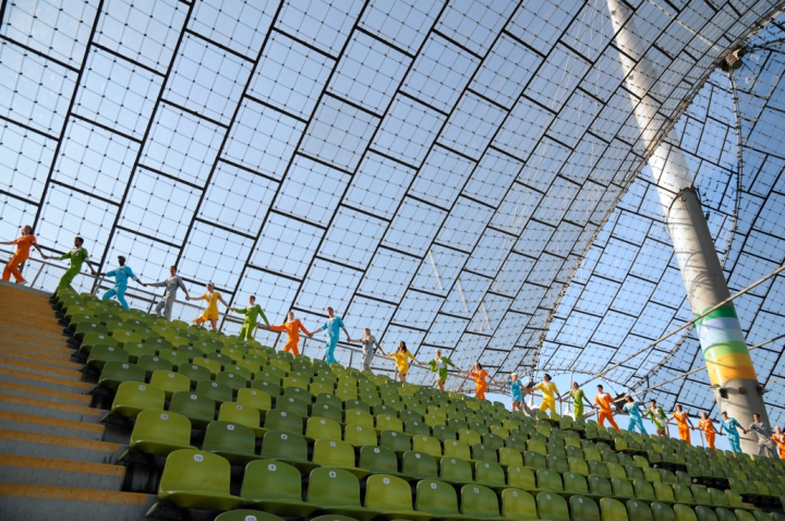 The picture is a photograph taken in the Olympic Stadium. Behind the last row of green chairs under the tent roof, the performers walk in a line, holding hands. Each performer wears a single-colour overall, ranging from orange and green to blue, silver and yellow.