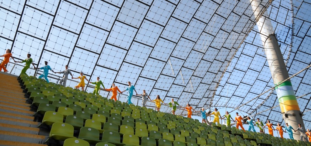 The picture is a photograph taken in the Olympic Stadium. Behind the last row of green chairs under the tent roof, the performers walk in a line, holding hands. Each performer wears a single-colour overall, ranging from orange and green to blue, silver and yellow.