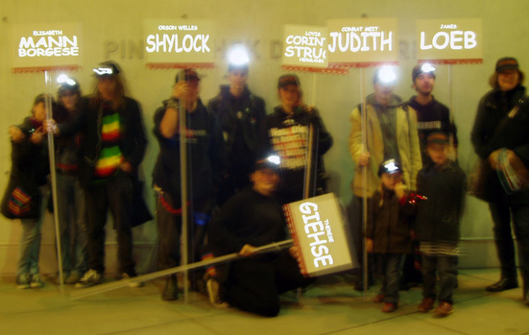 Die Fotografie zeigt eine Gruppe von Erwachsenen und Kindern, die dem Anschein nach in einer Art Unterführung stehen. Einige von ihnen tragen Stirnlampen und halten Schilder mit verschiedenen Namen und Texten, wie "James Loeb", "Orson Welles Shylock" oder "Elisabeth Mann Borgese" hoch.