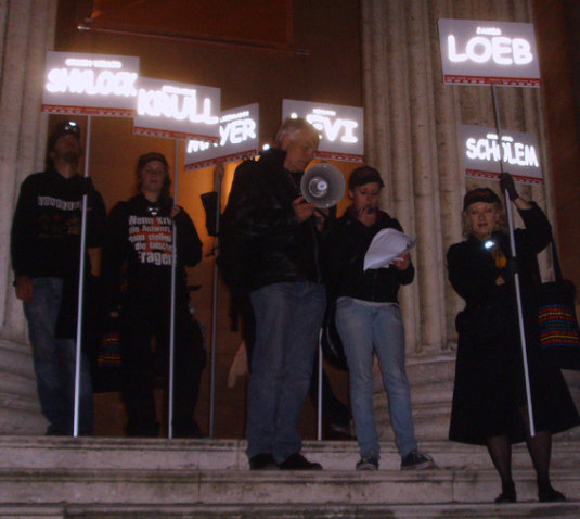 Photograph of a group of people standing on the steps of the Glyptothek on Königsplatz. It is evening. One man in the group is holding a megaphone, another participant is reading a text from a sheet of paper. The other participants on the steps hold up posters with different names, such as "Scholem", "Loeb" or "Levi", which glow in the dark.