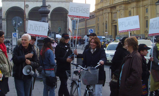 The photo shows a group of people standing at the taxi turnaround at Odeonsplatz. They are holding megaphones and placards. The white placards have names in capital letters such as 'Judith', 'Giehse', 'Pflaum' and 'Krull'. Passers-by on bicycles and on foot pass between them.