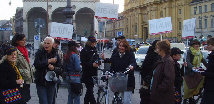 The photo shows a group of people standing at the taxi turnaround at Odeonsplatz. They are holding megaphones and placards. The white placards have names in capital letters such as 'Judith', 'Giehse', 'Pflaum' and 'Krull'. Passers-by on bicycles and on foot pass between them.