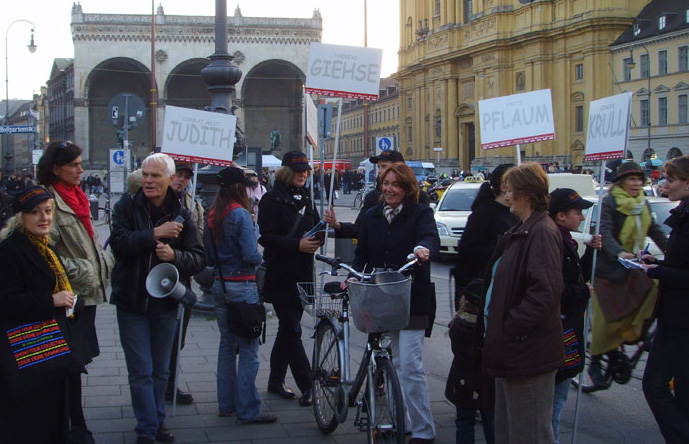 Die Fotografie zeigt eine Gruppe von Menschen, die an der Taxi-Wendeschleife beim Odeonsplatz stehen. Sie halten Megaphone und Plakate in der Hand. Auf den weißen Plakaten stehen Namen in Großbuchstaben wie "Judith", "Giehse", "Pflaum" und "Krull". Zwischen ihnen gehen und fahren Passant*innen mit Fahrrädern vorbei.