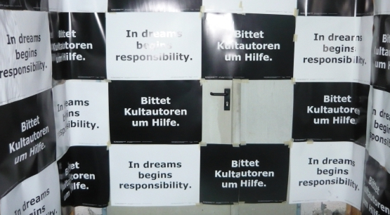 The photograph shows a large metal door covered with black and white rectangular pieces of paper in a chequered pattern. The text on all the black papers is written in white: "Ask cult authors for help". On all the white papers is written in black: "In dreams begins responsibility".
