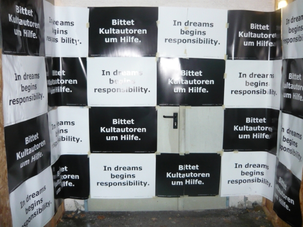 The photograph shows a large metal door covered with black and white rectangular pieces of paper in a chequered pattern. The text on all the black papers is written in white: "Ask cult authors for help". On all the white papers is written in black: "In dreams begins responsibility".