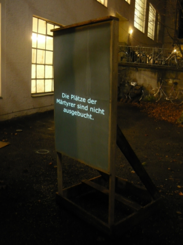 The photograph shows a wooden stand set up outdoors in a leafy backyard. In the background there is a warehouse with several bicycles parked in front of it. It is evening and the backyard and warehouse are lit. A black tarpaulin is stretched over the stand, onto which the text "Die Plätze der Märtyrer sind nicht ausgebucht." is projected.