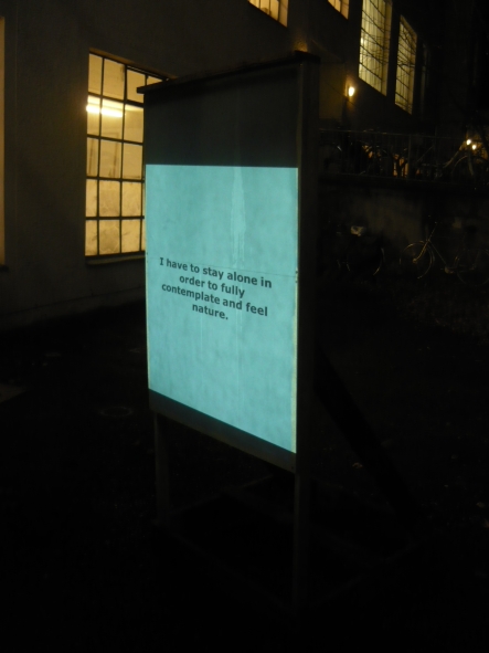 The photograph shows a wooden stand set up outside in a backyard. A warehouse can be seen in the background, illuminated from the inside. A black tarpaulin is stretched over the stand, onto which the text "I have to stay alone in order to fully contemplate and feel nature." is projected onto it.