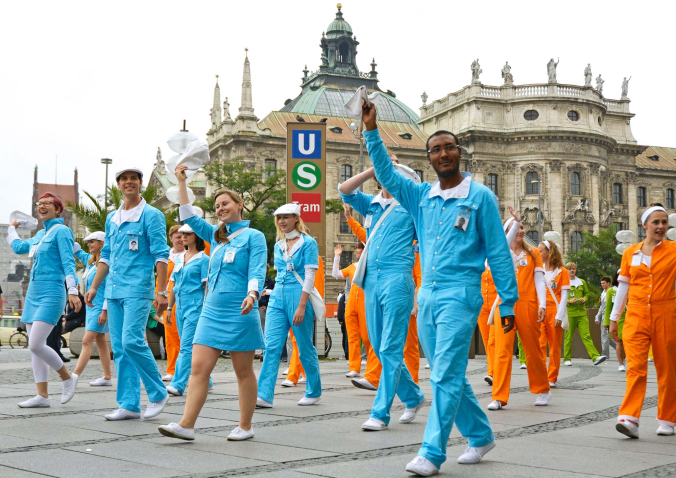 Das Bild zeigt eine Gruppe von Performer*innen in einfarbigen Overalls in den Farben Orange, Blau und Grün. Sie laufen auf dem Karlsplatz und winken umstehenden Passant*innen zu, die nicht im Bild zu sehen sind. Die Stimmung der Performer*innen scheint gelöst und fröhlich.